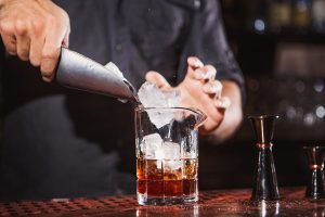Principle picture of male bartender dropping ice into a glass with liquid in the bottom