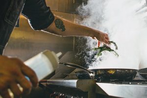 Principle man putting herbs into a cooking pan on stove with smoke coming out of it while holding bottle of sauce and has tattoo on arm