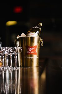 Principle medium shot of Miller High Life bucket of ice with Miller High Life bottle in it and four glasses sitting on bar