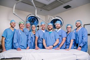 Kalamazoo Anesthesiology group of doctors and surgeons smiling at camera behind a surgey bed with two large lights on a swivel hanging over them